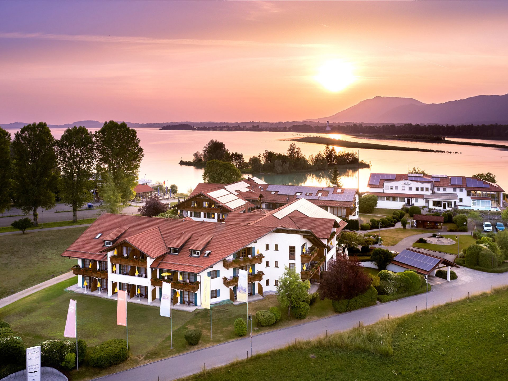 Wellnesshotel in Füssen im Allgäu | Hotel Sommer Hotelgebäude mit roten Dächern am Seeufer bei Sonnenuntergang
